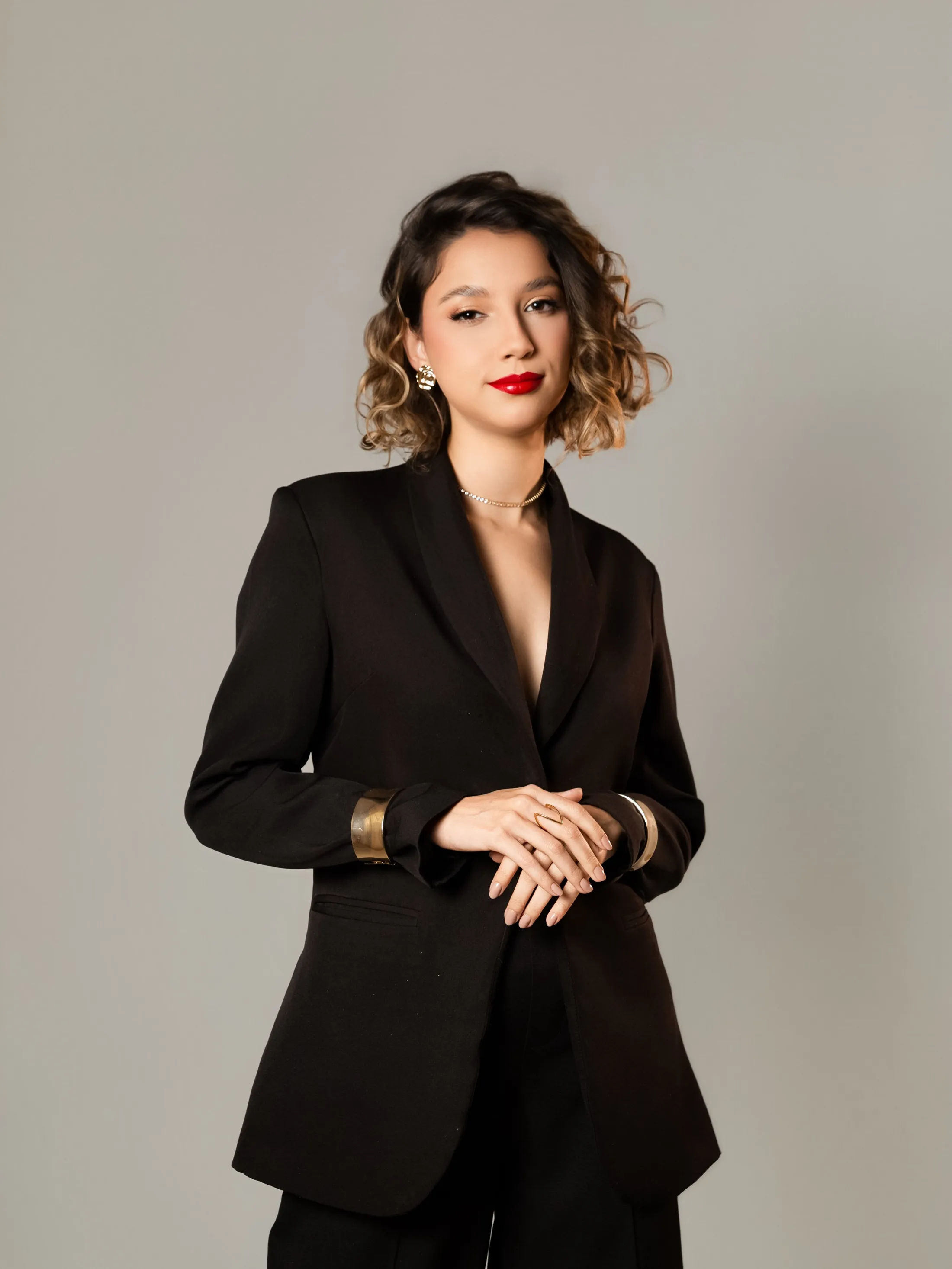 Mulher de terno preto em pé, com as mãos à frente do corpo.