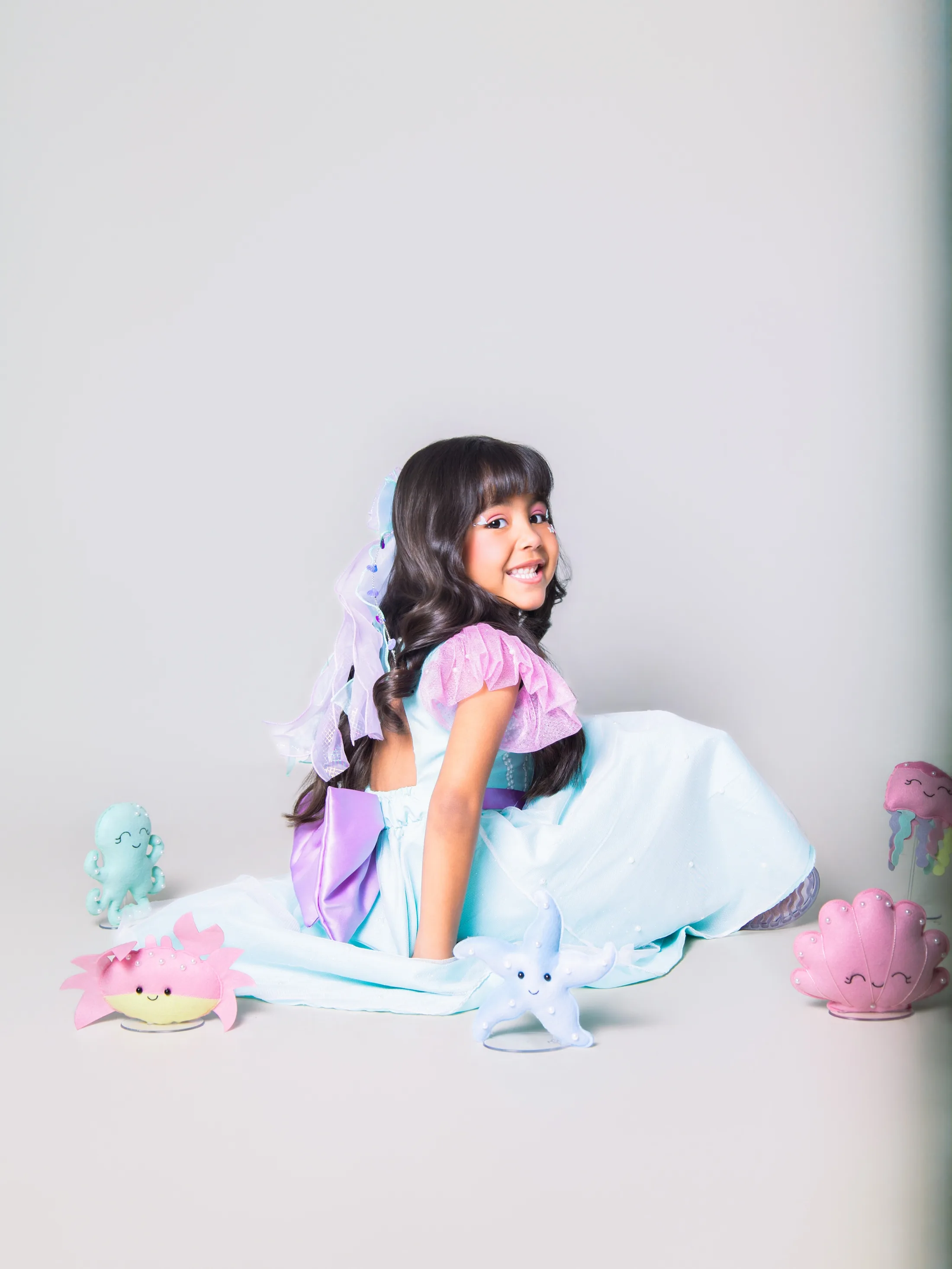 Menina de vestido azul e roxo sentada no chão do estúdio.