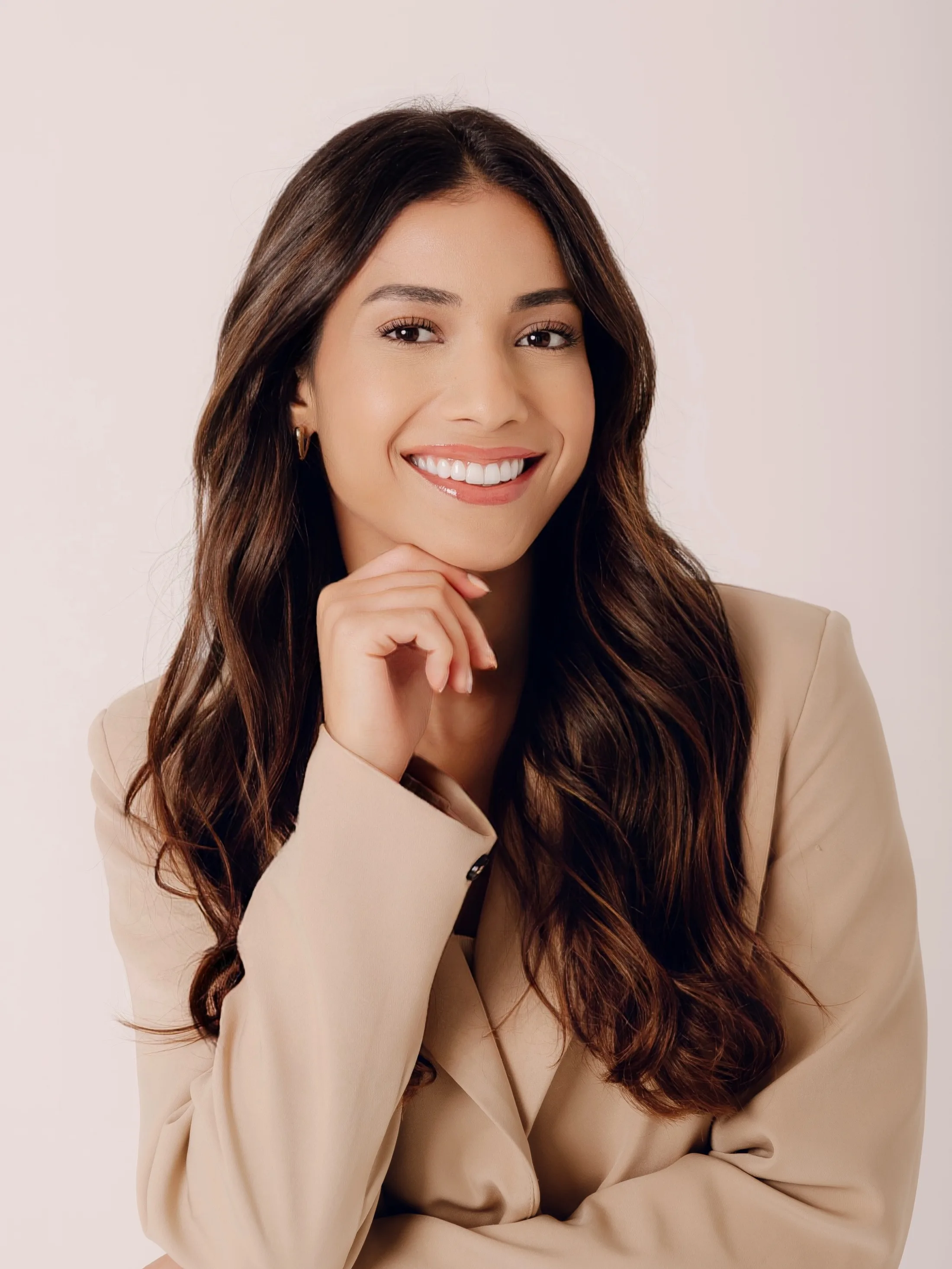 Mulher de blazer bege sorrindo com a mão junto ao rosto.