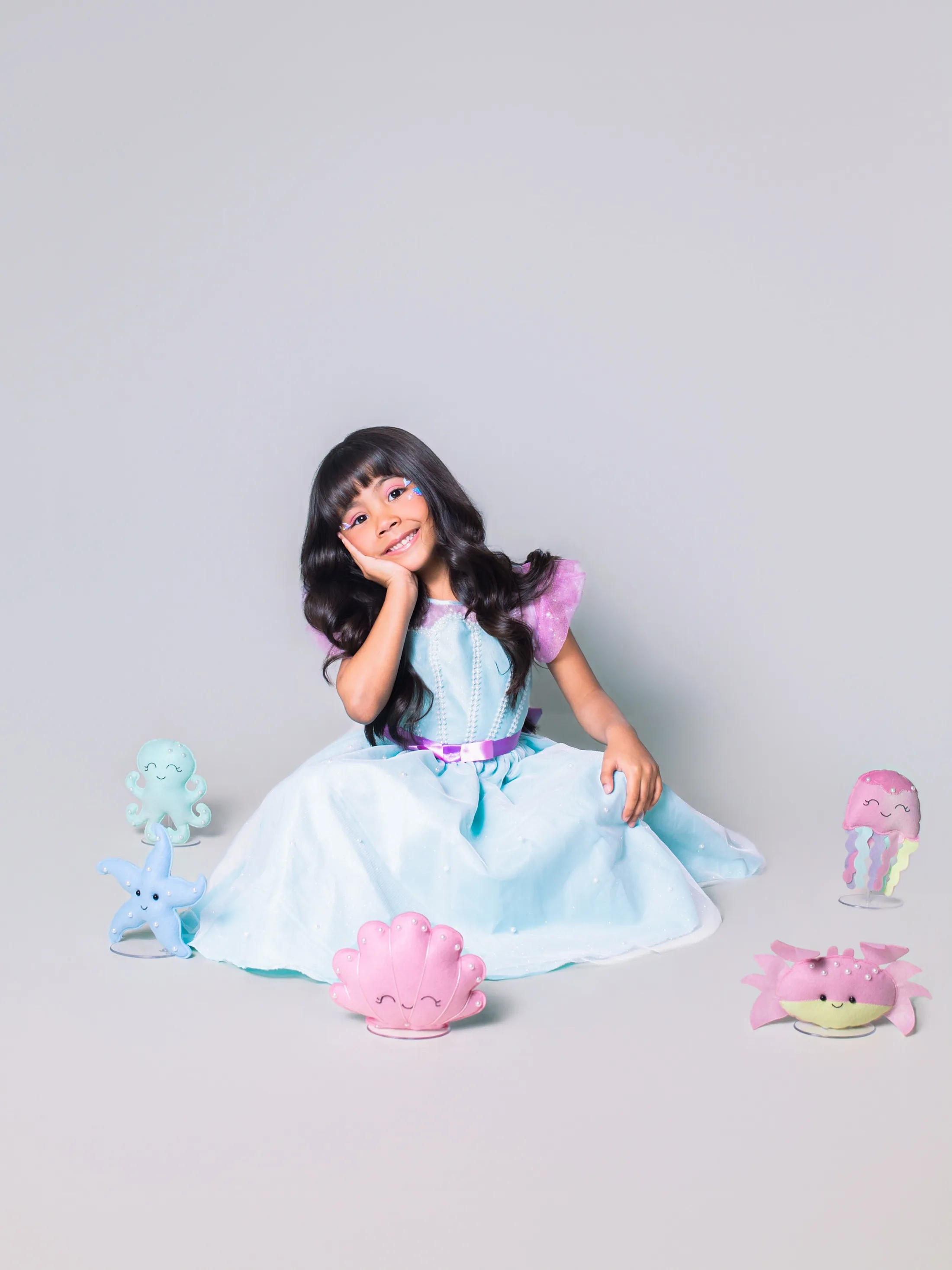 Menina de vestido azul e roxo sentada com a cabeça inclinada.