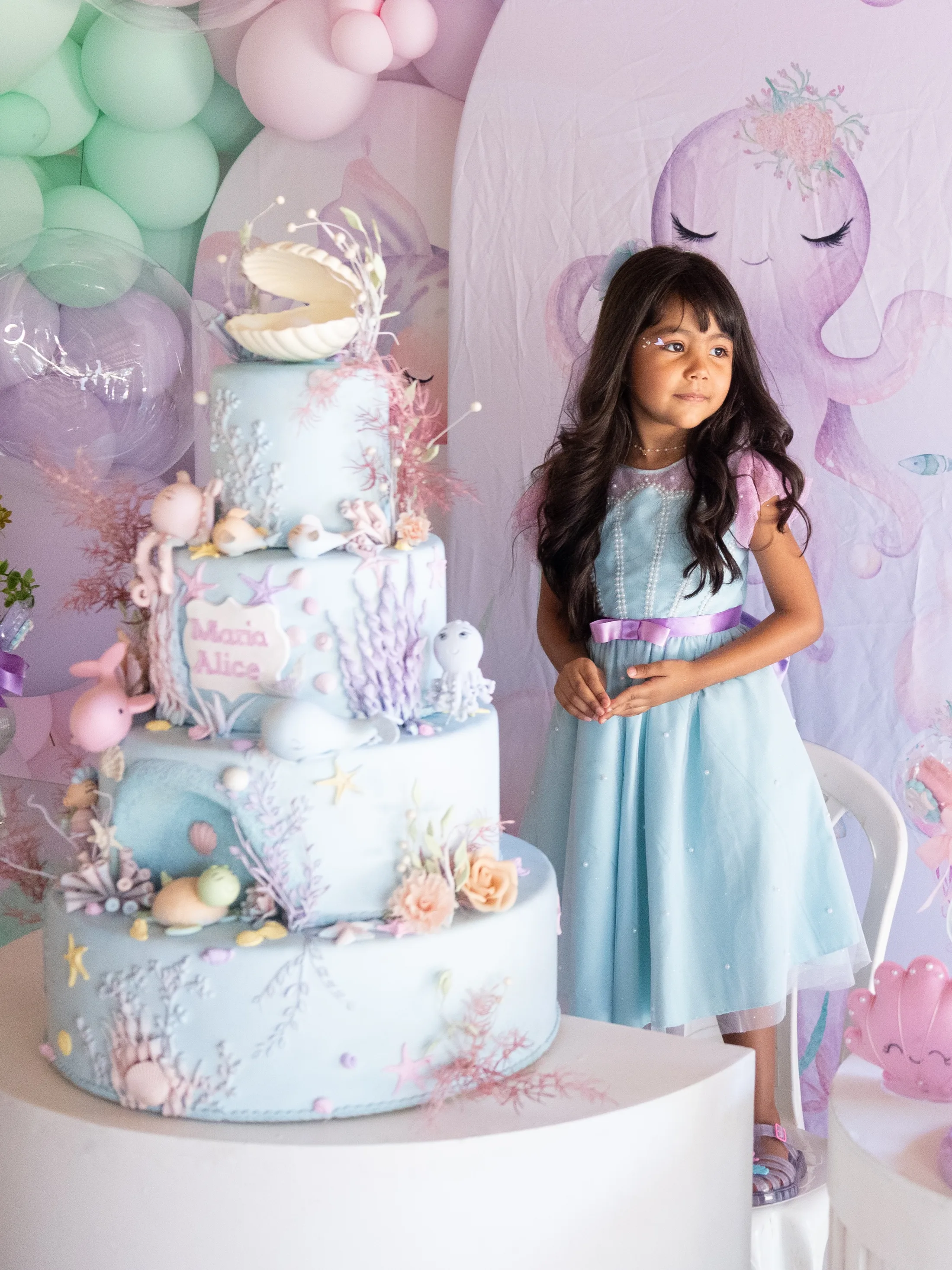 Menina de vestido azul ao lado de mesa de doces e balões.