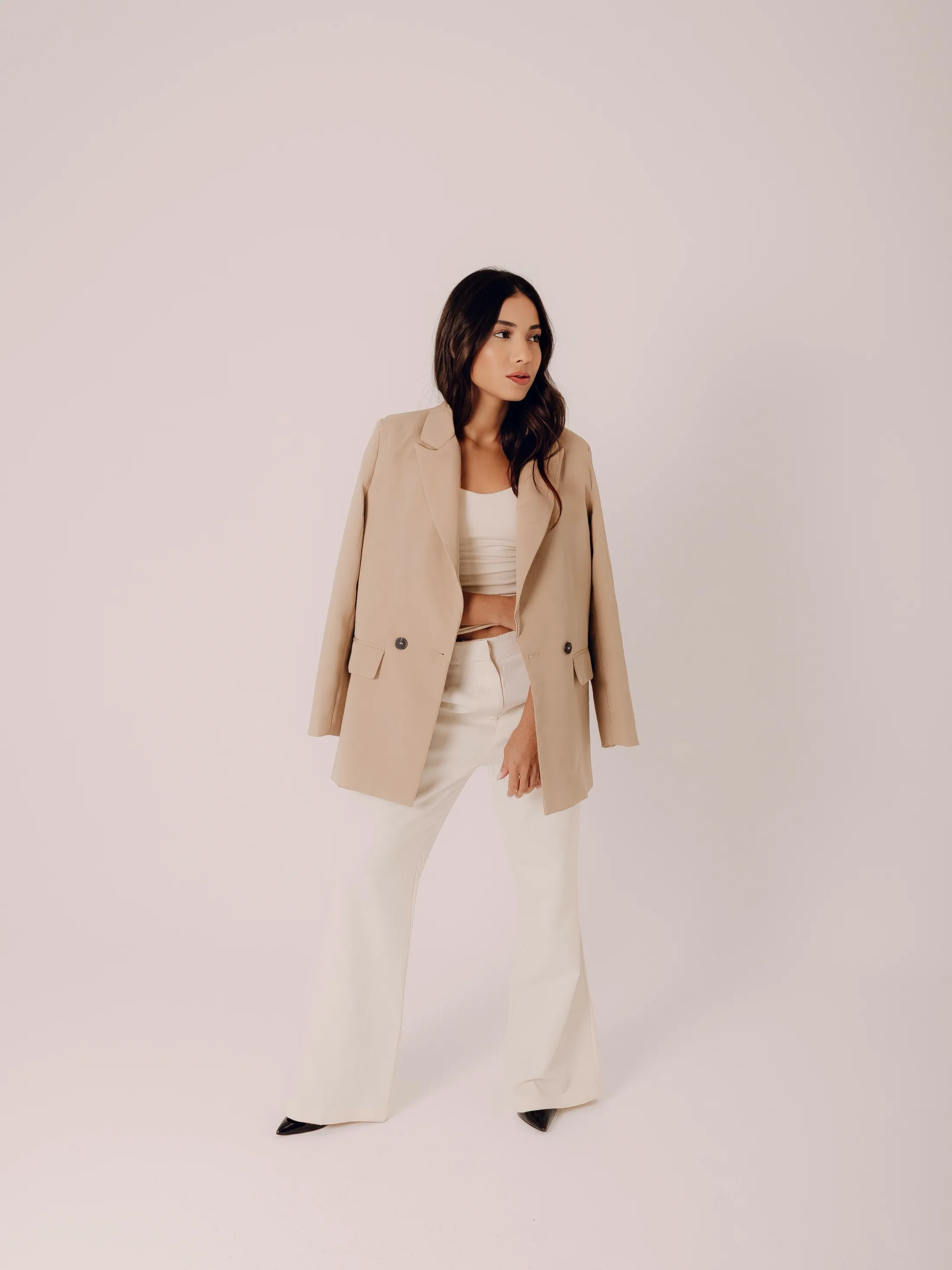 Mulher de blazer bege em pé em fundo claro.